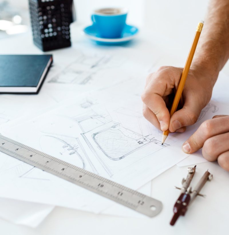 Picture of hands of young businessman holding pencil and drawing sketch at table. White modern office interior background.