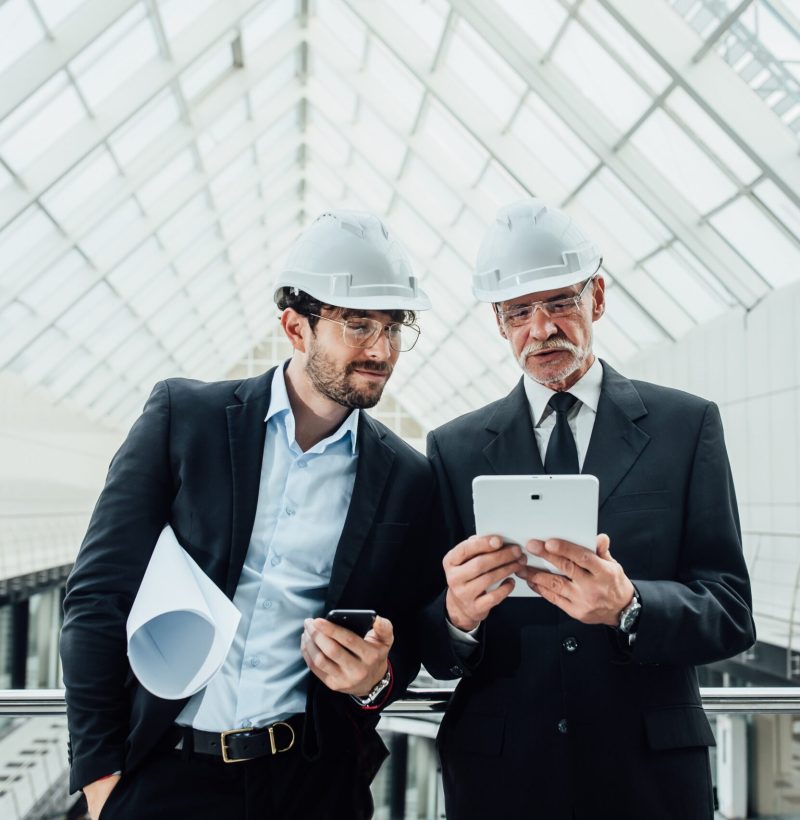 Two successful entrepreneur in helmet with project and laptop of new buildings stay near glass roof.